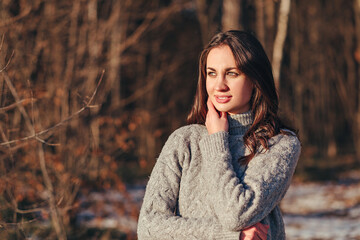 Portrait of pleasant young girl with dark hair in a warm knitted sweater against the background of forest. The rays of the sun fall beautifully on the woman. A look at the landscape.