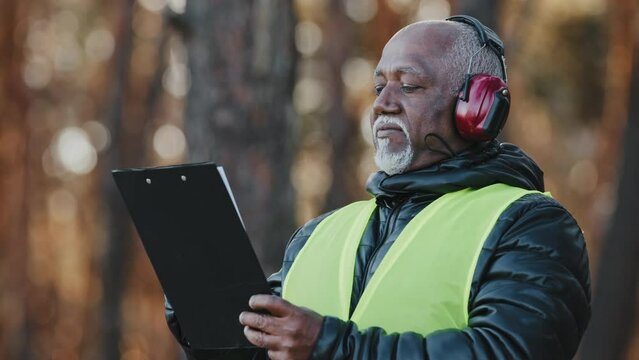 Close-up african american male professional foreman forestry engineer standing outdoors wearing protective headphones assessing situation making tree felling plan writing data reforestation projectt