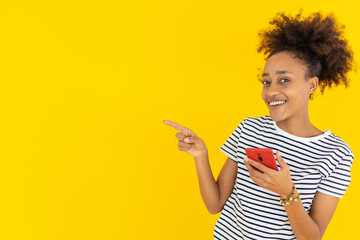 Smiling young african woman with smartphone pointing with finger empty space isolated yellow color background