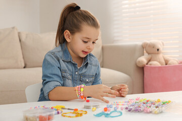 Cute little girl making beaded jewelry at table in room