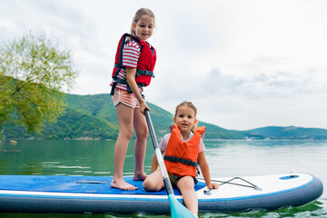 Girls padding on stand up paddle boarding on lake district. Children in swim life vest learning paddleboarding on SUP board. Family active leisure and local getaway with kids concept - obrazy, fototapety, plakaty
