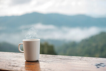 White mug of hot coffee or tea on wooden table in the morning with mountain and nature background