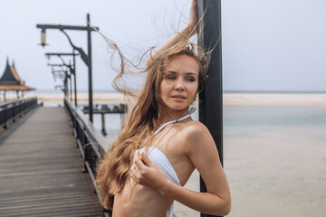 A young beautiful woman with brown hair in a white bikini elegantly poses on the pier on a beach background with sand, ocean and sky. Tropical photo shoot.