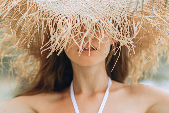 A close-up picture with a young cute girl with brown hair in a white swimsuit and a straw hat covering her face. Blurred background. Mysterious posing.