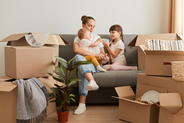 Pretty young mother and her cute little daughters sitting on cough of new apartment and unpacking moving boxes, expressing positive emotions, surrounded with packages with belongings.