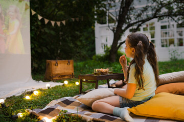 Family summer outdoor movie night. Girl sitting on blanket and pillows, eating homemade popcorn and watching film on DIY screen with from projector. Summer outdoor weekend activities with kids.
