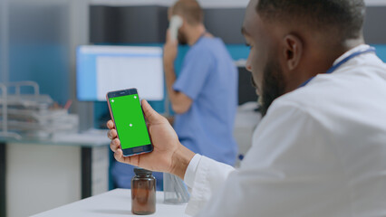 African american therapist doctor holding mock up green screen chroma key smartphone with isolated display. Physician man analyzing disease report working at medical expertise in hospital office