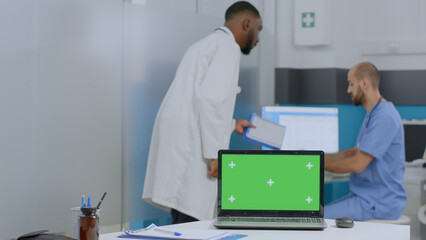Mock up green screen chroma key laptop computer with isolated display standing on table in hospital office. Multi-ethnic team discussing disease symptoms working at healthcare treatment