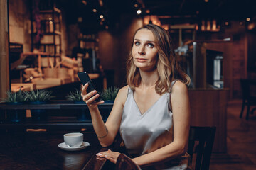 Portrait of serious focused pensive model sitting and posing at the table of an expensive restaurant and holding a smartphone and looking away. Working moment concept