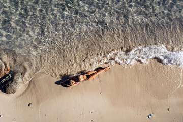 Overhead view of a sexy slim and attractive model in a black bikini lying on the wet sand of the beach and the waves of the sea hitting her tanned body. Perfect vacation concept