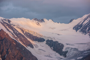 Dramatic aerial view to high snow mountain range in early morning at dawn. Awesome scenery with sunlit snow mountains in cloudy sky at sunrise. Scenic landscape with large glacier in sunrise colors.
