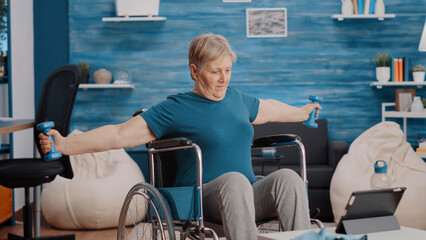 Aged woman stretching arms muscles with dumbbells, sitting in wheelchair and watching video of...