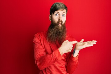 Redhead man with long beard pointing open palm with finger making fish face with mouth and squinting eyes, crazy and comical.