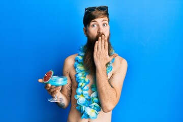 Redhead man with long beard wearing swimsuit drinking tropical cocktail covering mouth with hand,...