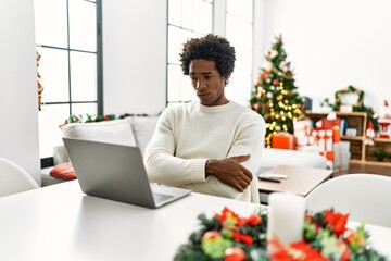 Young african american man using laptop sitting on the table by christmas tree skeptic and nervous,...