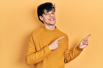 Handsome hipster young man wearing casual yellow sweater smiling and looking at the camera pointing with two hands and fingers to the side.