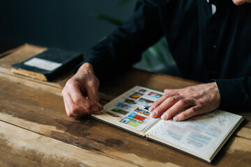 Close-up of unrecognizable senior adult man inserting postage stamps at collection book sitting at wooden table, selective focus. Mature aged man looking album with old stamps philately at home.