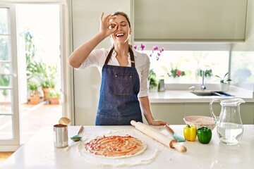 Beautiful blonde woman wearing apron cooking pizza smiling happy doing ok sign with hand on eye looking through fingers