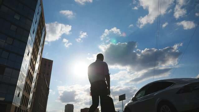 Young businessman in formal wear going to his modern electric car. Successful entrepreneur with briefcase opening door and getting inside to new electrical SUV vehicle. Cityscape at background