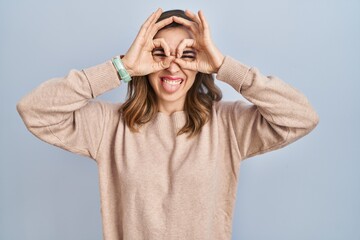 Young woman standing over isolated background doing ok gesture like binoculars sticking tongue out, eyes looking through fingers. crazy expression.