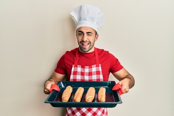 Young hispanic man wearing baker uniform holding homemade bread winking looking at the camera with...