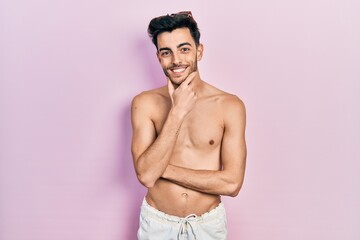 Fototapeta premium Young hispanic man wearing swimwear shirtless looking confident at the camera smiling with crossed arms and hand raised on chin. thinking positive.
