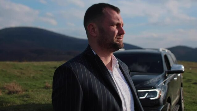 A man in a business suit with a serious expression stands on a background of mountains near a black SUV. Businessman near the car