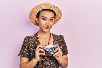 Beautiful hispanic woman with short hair wearing summer hat holding vintage camera relaxed with serious expression on face. simple and natural looking at the camera.