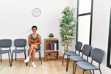 Young hispanic woman reading book sitting on chair at waiting room