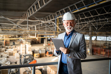 Successful factory manager or CEO holding tablet computer and checking production results in industrial plant while workers and forklifts working in background. - obrazy, fototapety, plakaty