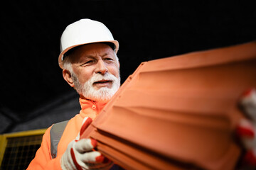 Construction worker in safety uniform and hardhat holding roof tiles at construction site.