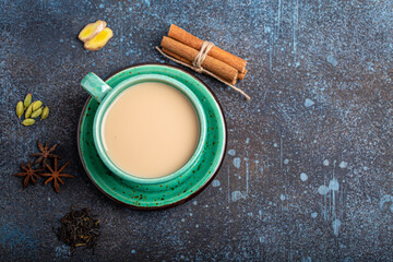 Healthy Indian beverage masala chai - tea hot drink with milk and spices in rustic green teacup with ingredients for making masala chai from above on concrete blue background copy space