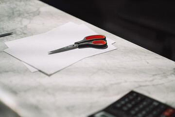 Photo of scissors with red handles on a table with calculator paper and a pen.