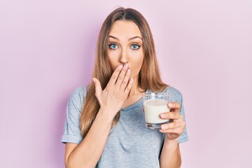 Young blonde girl holding glass of milk covering mouth with hand, shocked and afraid for mistake....