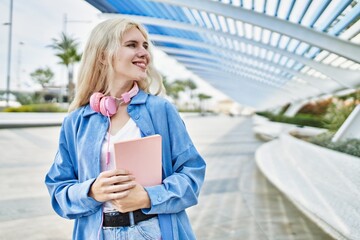 Fototapeta premium Beautiful blonde young woman smiling happy holding book and wearing headphones outdoors on a sunny day