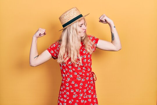 Beautiful caucasian woman with blond hair wearing summer hat showing arms muscles smiling proud. fitness concept.