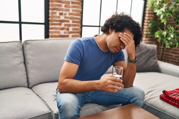 Young hispanic man unwell drinking water sitting on sofa at home