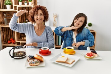 Family of mother and down syndrome daughter sitting at home eating breakfast gesturing with hands showing big and large size sign, measure symbol. smiling looking at the camera. measuring concept.