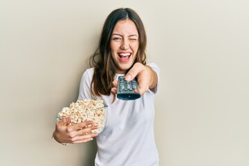 Young brunette woman eating popcorn using tv control winking looking at the camera with sexy expression, cheerful and happy face.