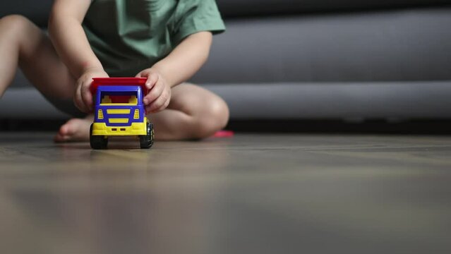unrecognizable baby boy kid in green shirt driving colorful plastic toy car truck towards camera on brown wooden floor home living room. concept of children indoor activity or transport construction 