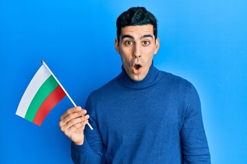 Handsome hispanic man holding bulgarian flag scared and amazed with open mouth for surprise, disbelief face - obrazy, fototapety, plakaty