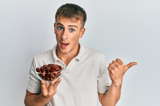 Young caucasian man holding bowl with dates pointing thumb up to the side smiling happy with open mouth