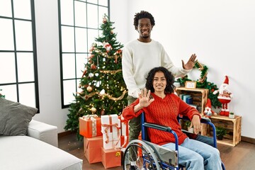Young interracial couple with woman sitting on wheelchair by christmas tree showing and pointing up with fingers number ten while smiling confident and happy.