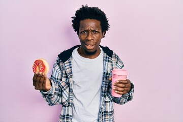 Young african american man eating doughnut and drinking coffee clueless and confused expression....