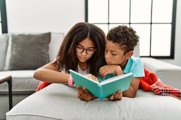 Two siblings lying on the sofa reading a book checking the time on wrist watch, relaxed and...