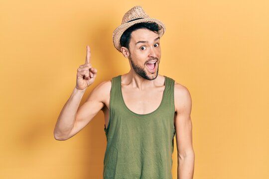 Young hispanic man wearing summer hat pointing finger up with successful idea. exited and happy. number one.