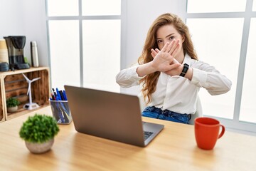Young caucasian woman working at the office using computer laptop rejection expression crossing...