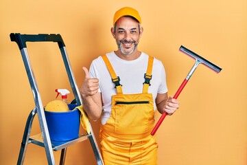 Handsome middle age man with grey hair wearing glass cleaner uniform and squeegee smiling happy and positive, thumb up doing excellent and approval sign