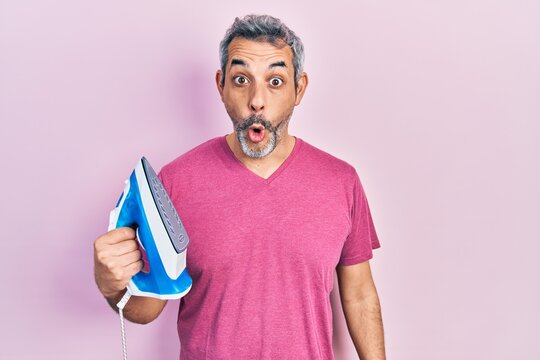 Handsome middle age man with grey hair holding electric steam iron scared and amazed with open mouth for surprise, disbelief face