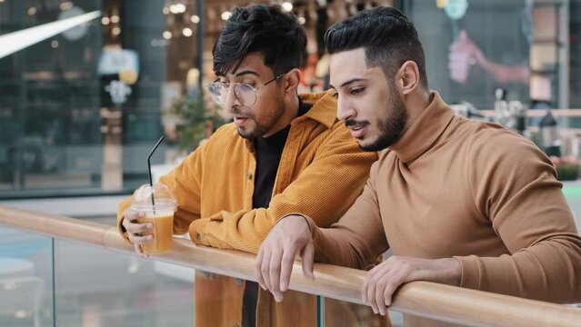 Two positive arab guys friends students stand in public place in shopping and entertainment center talking communicate pointing fingers choose where can have good time enjoy weekend man drinking juice